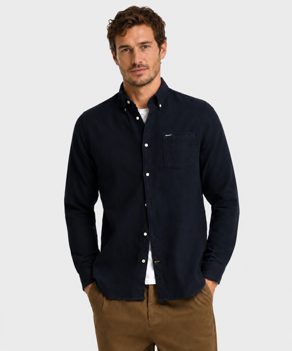 A man wearing a Barbour dark navy blue herringbone long-sleeve button-down shirt with a chest pocket, paired with brown pants, standing against a light gray background.