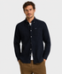 A man wearing a Barbour dark navy blue herringbone long-sleeve button-down shirt with a chest pocket, paired with brown pants, standing against a light gray background.