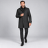 A man modeling a dark charcoal gray wool-blend coat over a black zip-up sweater, black trousers, and black dress shoes, walking against a light gray studio background.