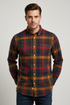 A man wearing a long-sleeve button-down plaid shirt in red, orange, and dark green tones, paired with dark blue jeans, photographed against a neutral gray studio background.