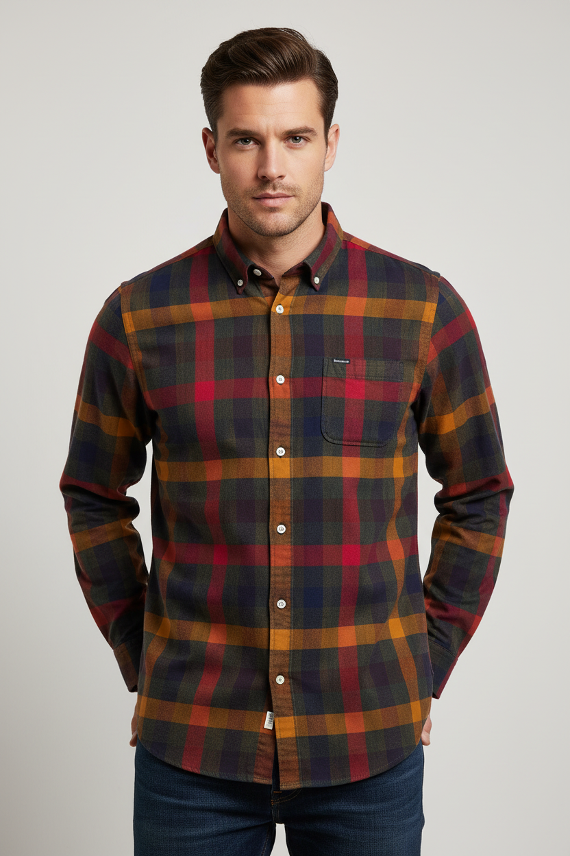 A man wearing a long-sleeve button-down plaid shirt in red, orange, and dark green tones, paired with dark blue jeans, photographed against a neutral gray studio background.