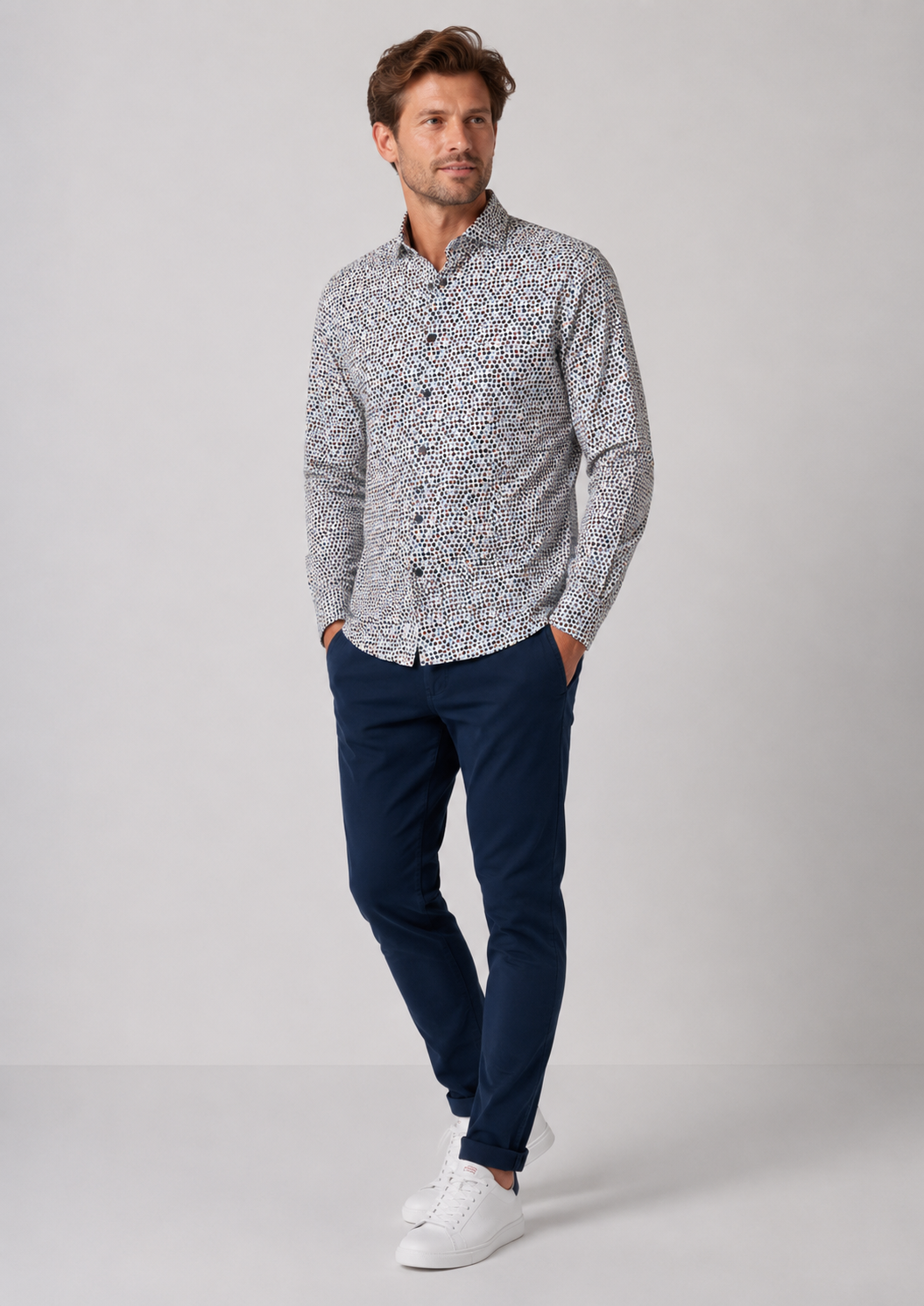 A man modeling a long-sleeve button-up shirt with a dense black and white abstract pattern, paired with navy blue trousers and white sneakers, against a light gray studio background.