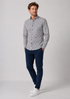 A man modeling a long-sleeve button-up shirt with a dense black and white abstract pattern, paired with navy blue trousers and white sneakers, against a light gray studio background.