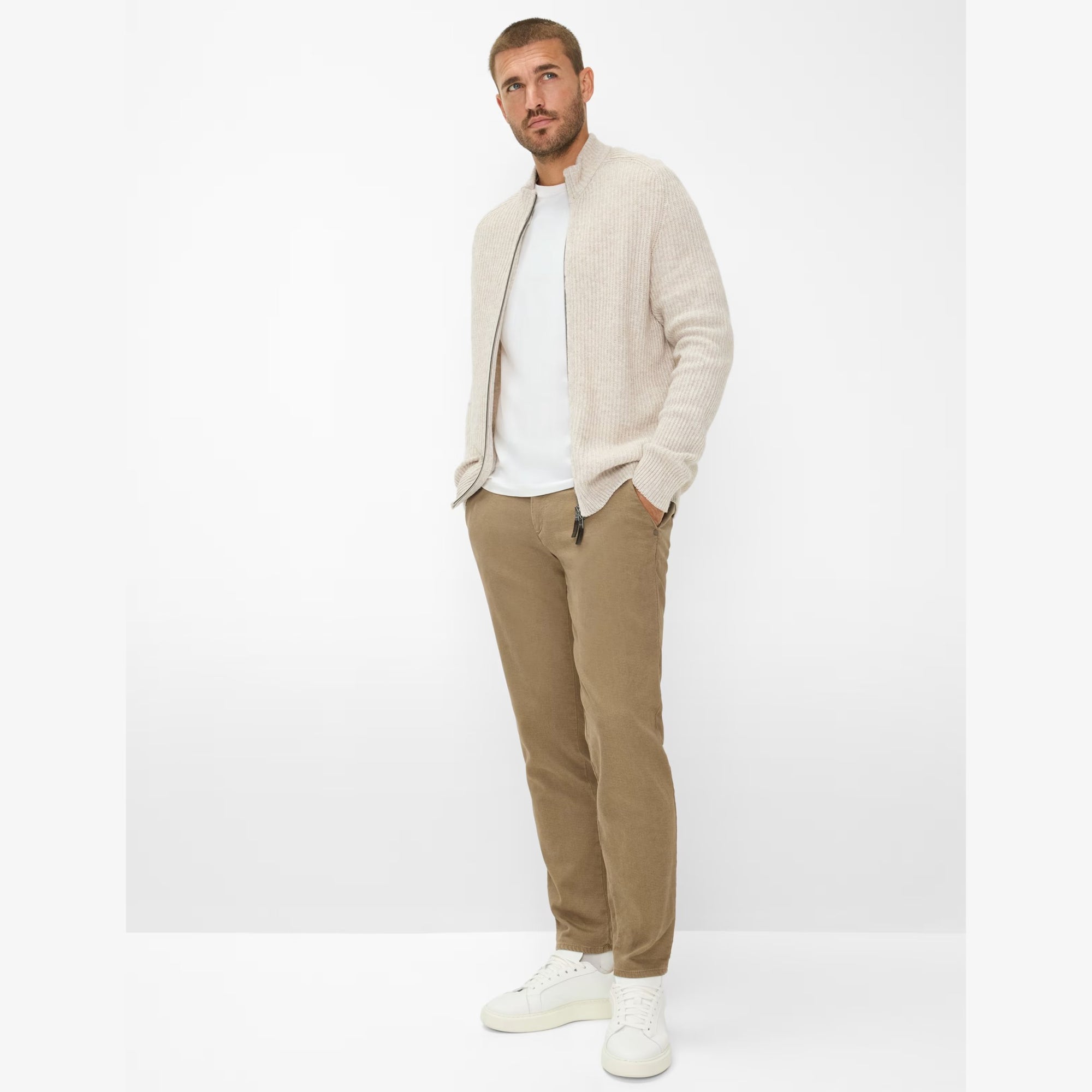 Man wearing a beige cardigan, white shirt, beige pants, and white sneakers on a white background
