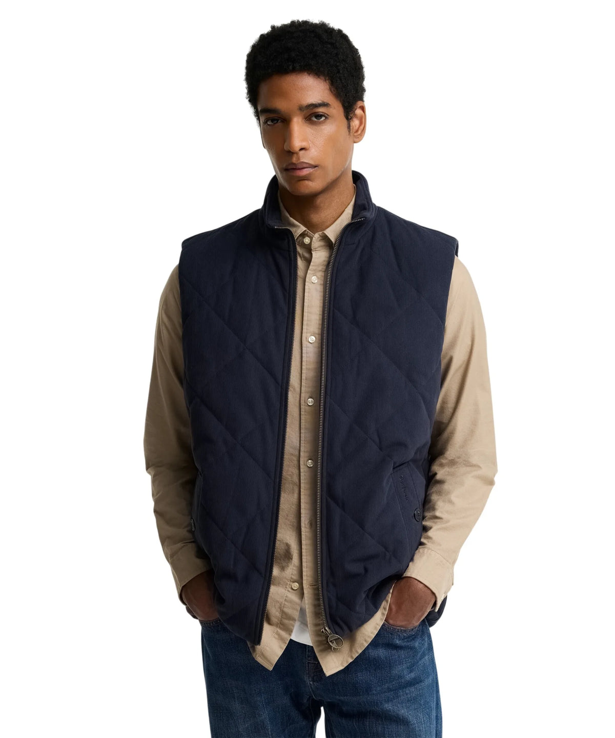 A man wearing a Barbour navy blue diamond-quilted zip-up gilet over a beige button-down shirt and blue jeans, photographed against a solid white background.