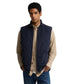 A man wearing a Barbour navy blue diamond-quilted zip-up gilet over a beige button-down shirt and blue jeans, photographed against a solid white background.