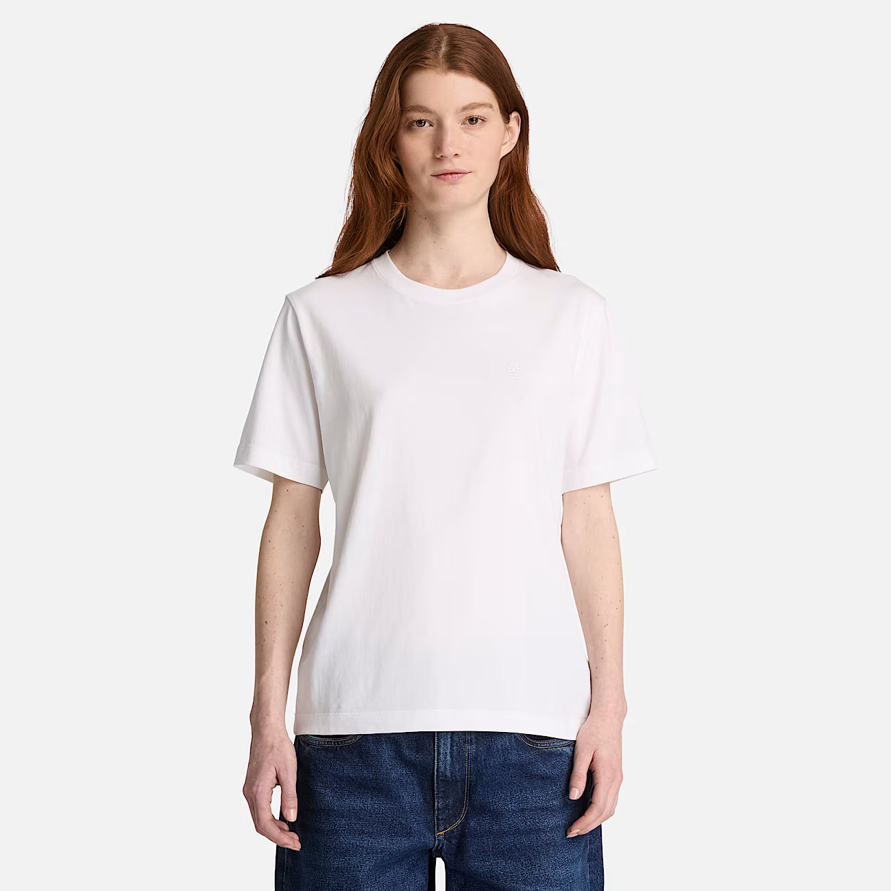 A woman wearing a Timberland plain white short-sleeve crewneck t-shirt and dark blue jeans, standing against a light gray studio background.