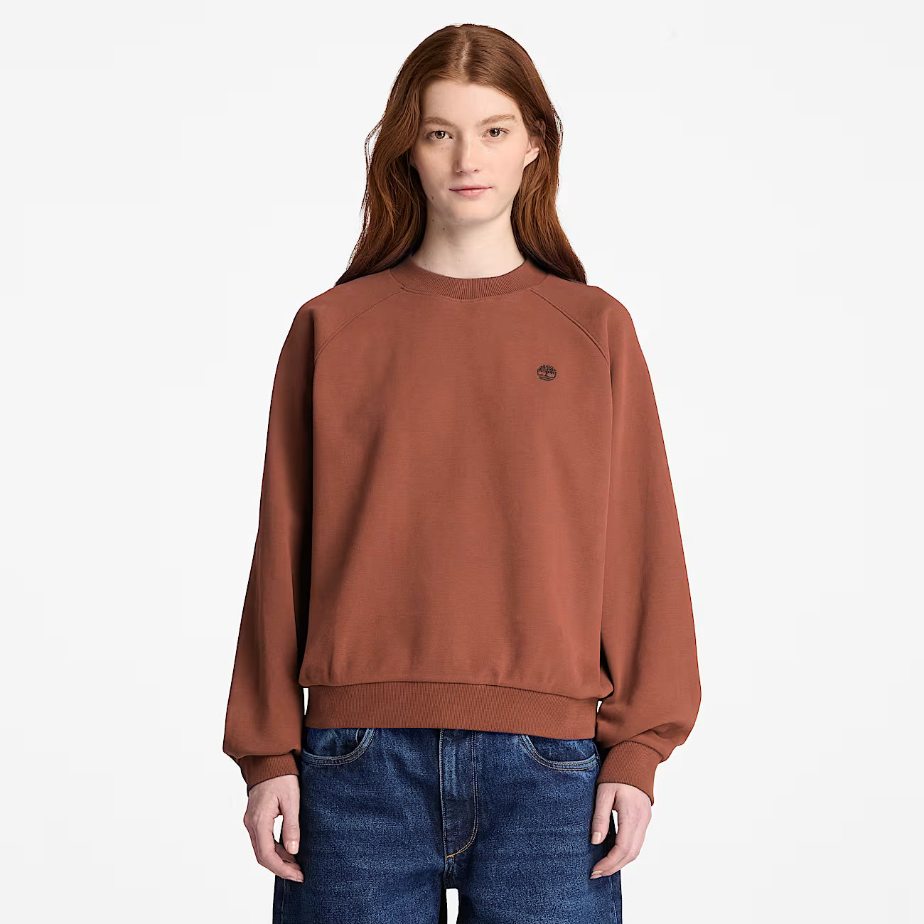 A woman modeling a Timberland rust-brown crewneck sweatshirt with a small embroidered logo on the left chest, paired with blue jeans, against a plain white studio background.