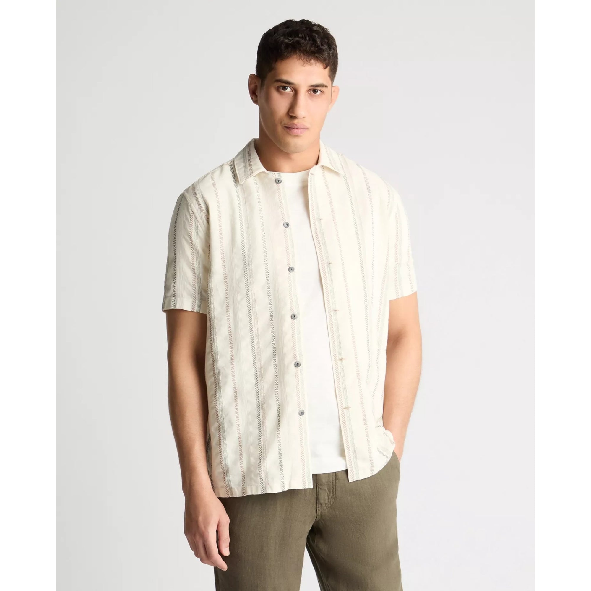 Man wearing a white striped shirt over a white t-shirt with green pants on a light gray background