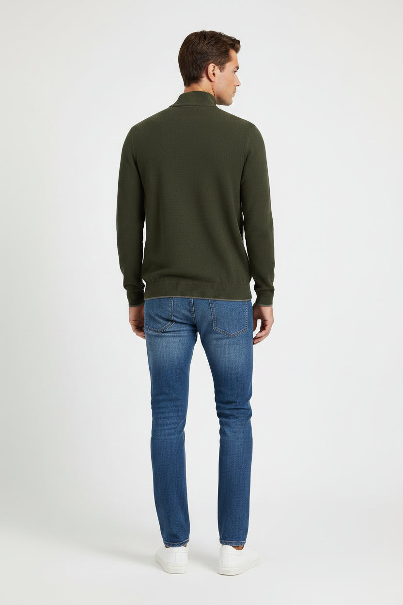 A man modeling a dark olive green long-sleeve quarter-placket sweater with blue jeans and white sneakers, standing against a light gray studio background.