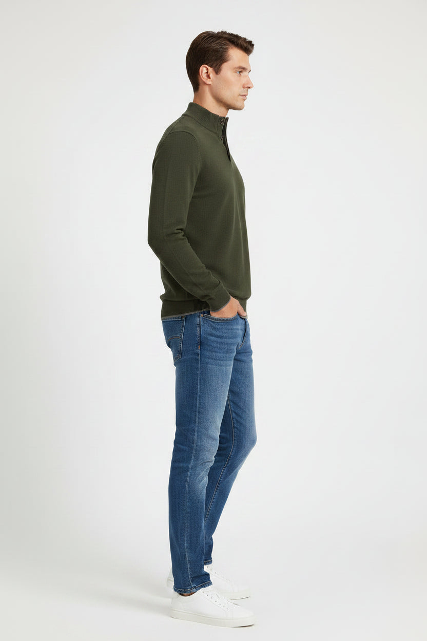 A man modeling a dark olive green long-sleeve quarter-placket sweater with blue jeans and white sneakers, standing against a light gray studio background.