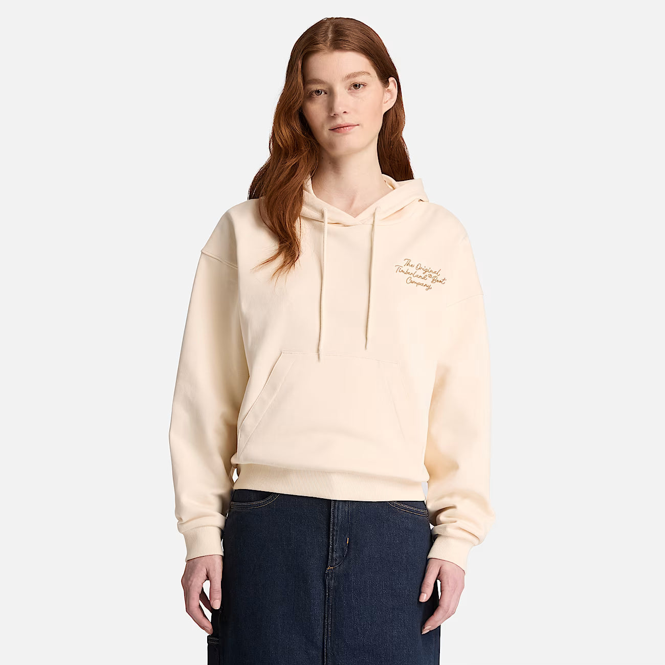 A woman modeling a cream-colored Timberland pullover hoodie with a small embroidered logo on the left chest, paired with dark blue jeans, against a light gray studio background.