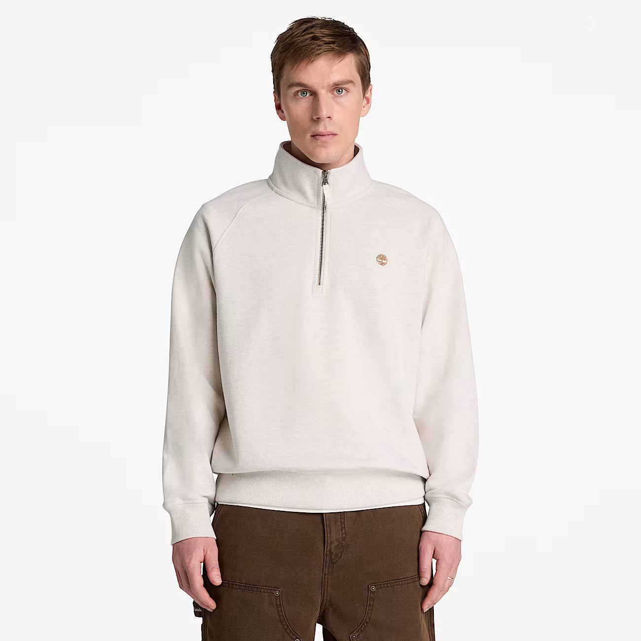 A man wearing a cream-colored Timberland quarter-zip pullover sweater with a small embroidered logo on the left chest, paired with brown pants, against a plain white background.
