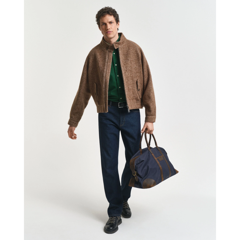Man wearing a GANT brown jacket and blue jeans, holding a navy duffel bag on a white background