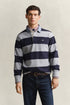 A man wearing a navy and light gray GANT horizontally striped long-sleeve rugby shirt over a collared shirt and jeans, standing against a plain beige studio background.