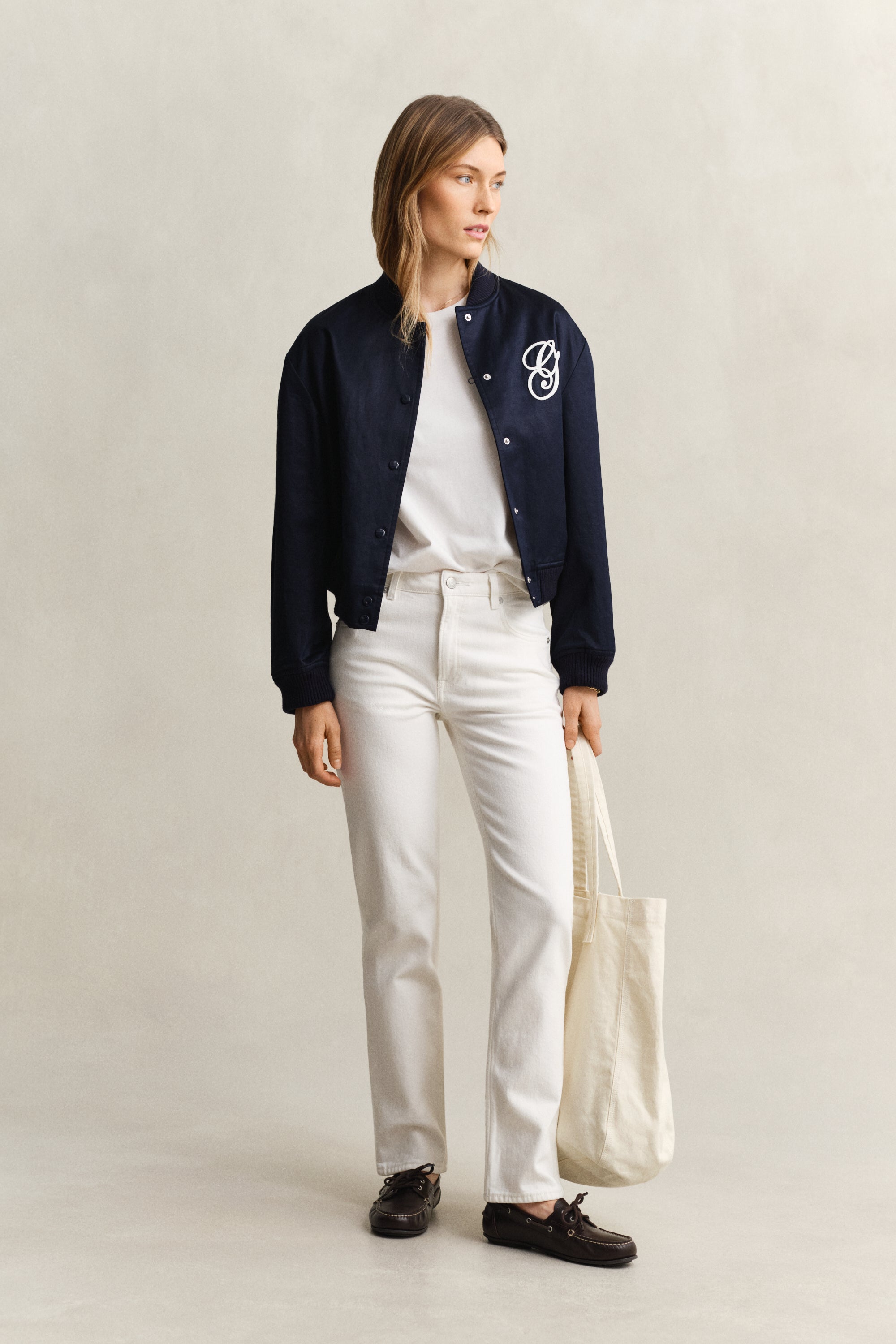 Person wearing a navy bomber jacket with a logo, white shirt, and white pants on a beige background