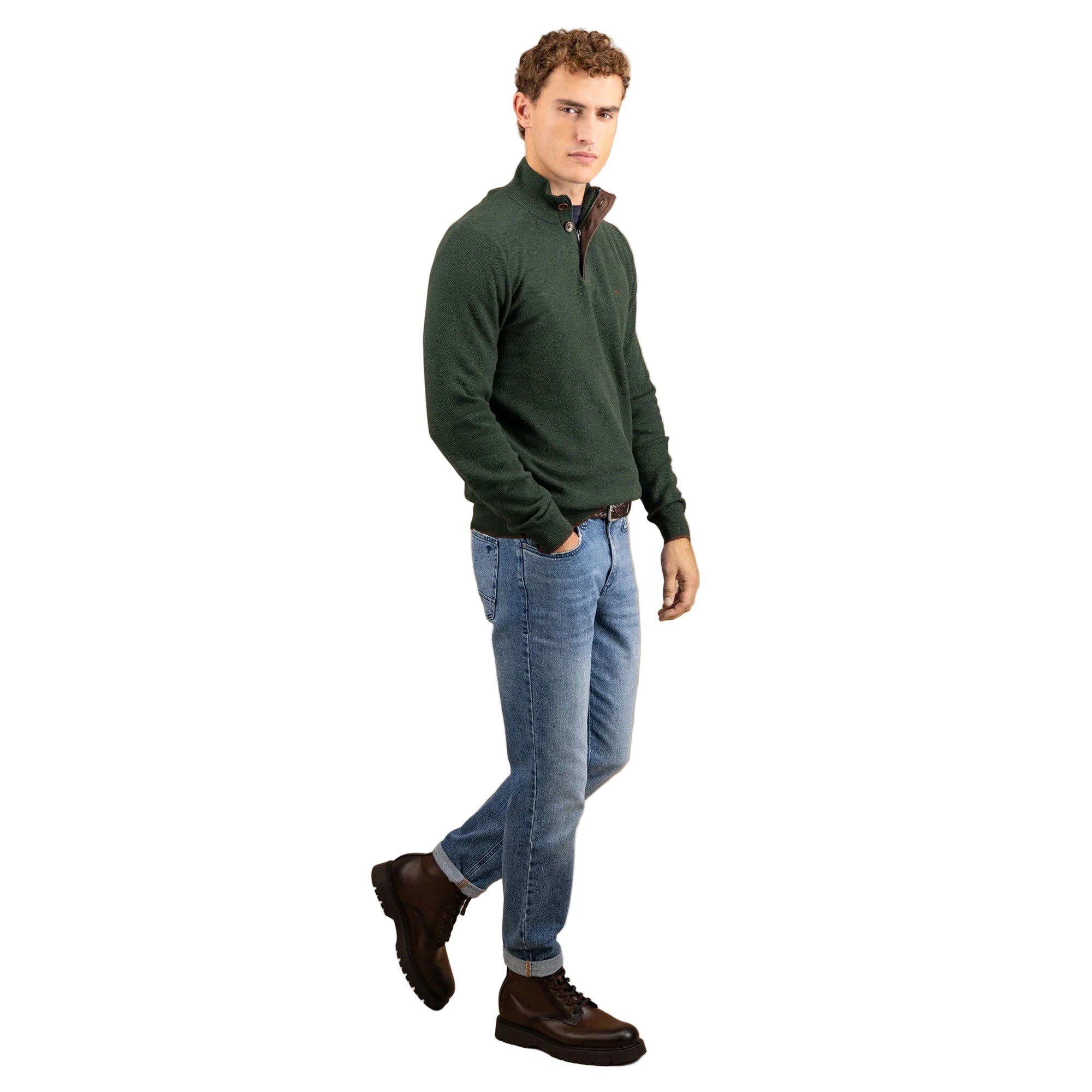 Man wearing a green sweater, blue jeans, and brown boots on a beige background