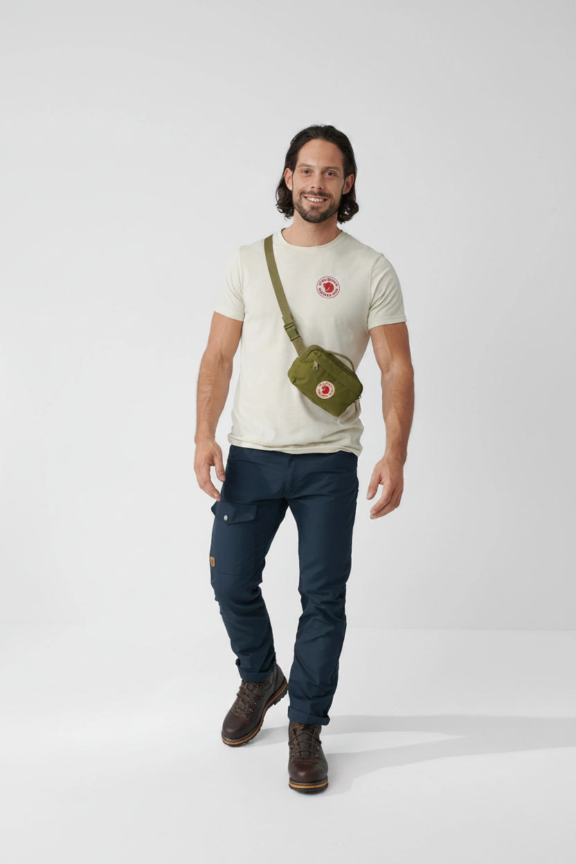 Man wearing a white t-shirt, dark pants, and brown boots with a green crossbody bag on a light gray background