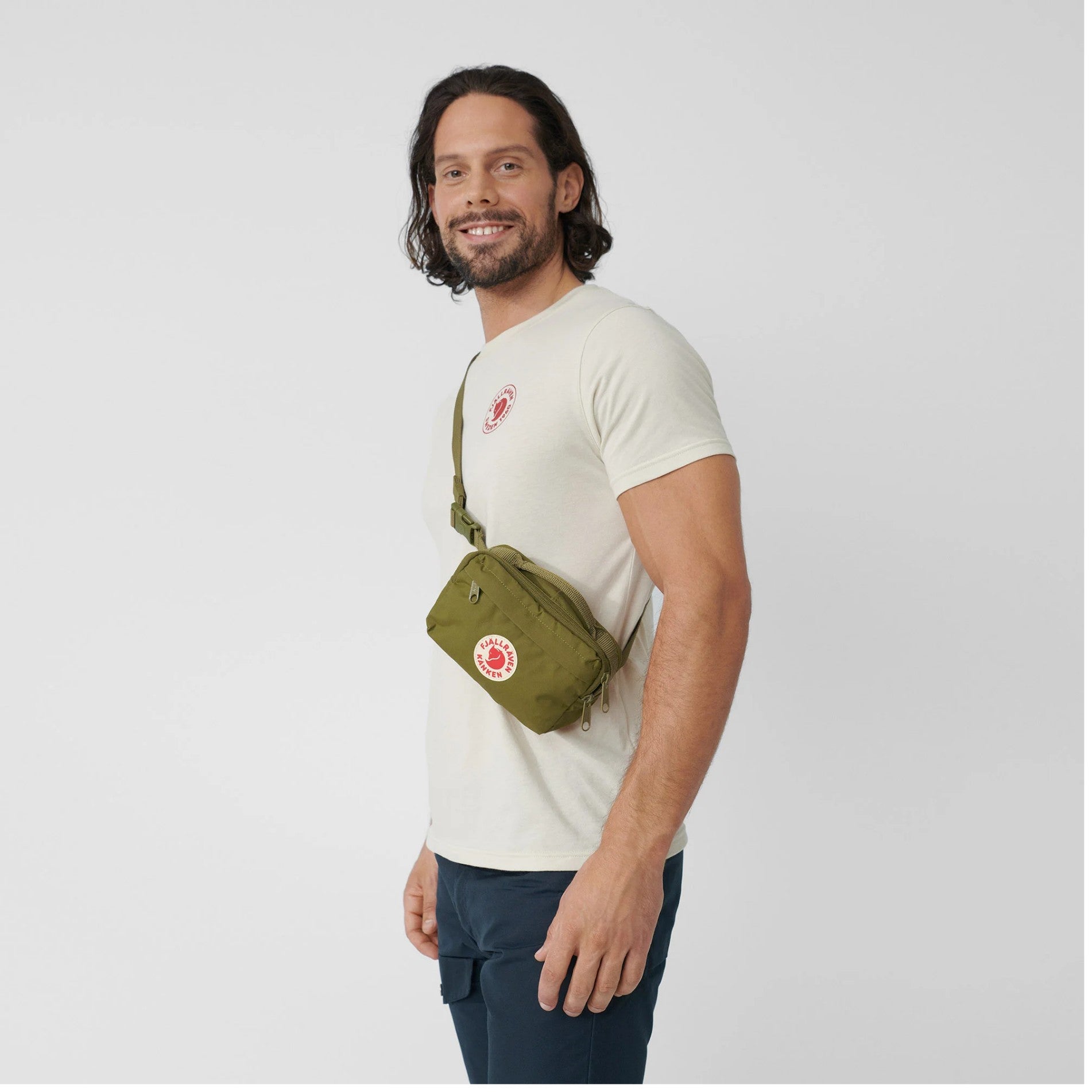 Man wearing a white t-shirt and green crossbody bag on a light gray background