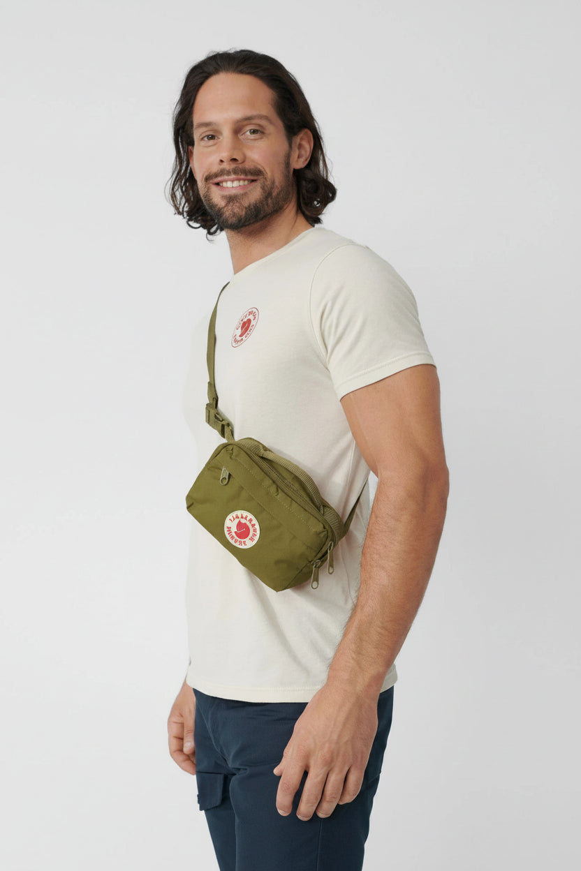 Man wearing a white t-shirt and green crossbody bag on a light gray background