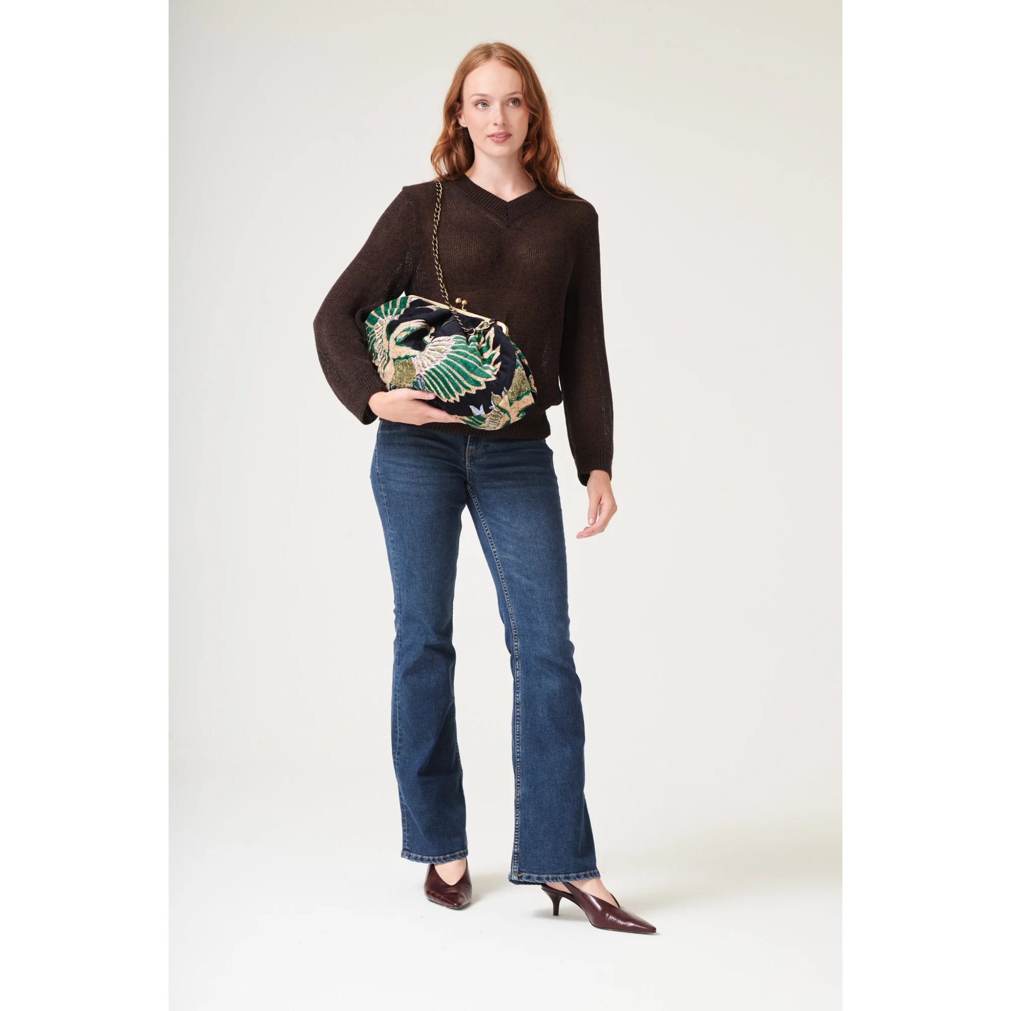 Woman wearing a brown sweater and blue jeans, holding a patterned handbag on a white background