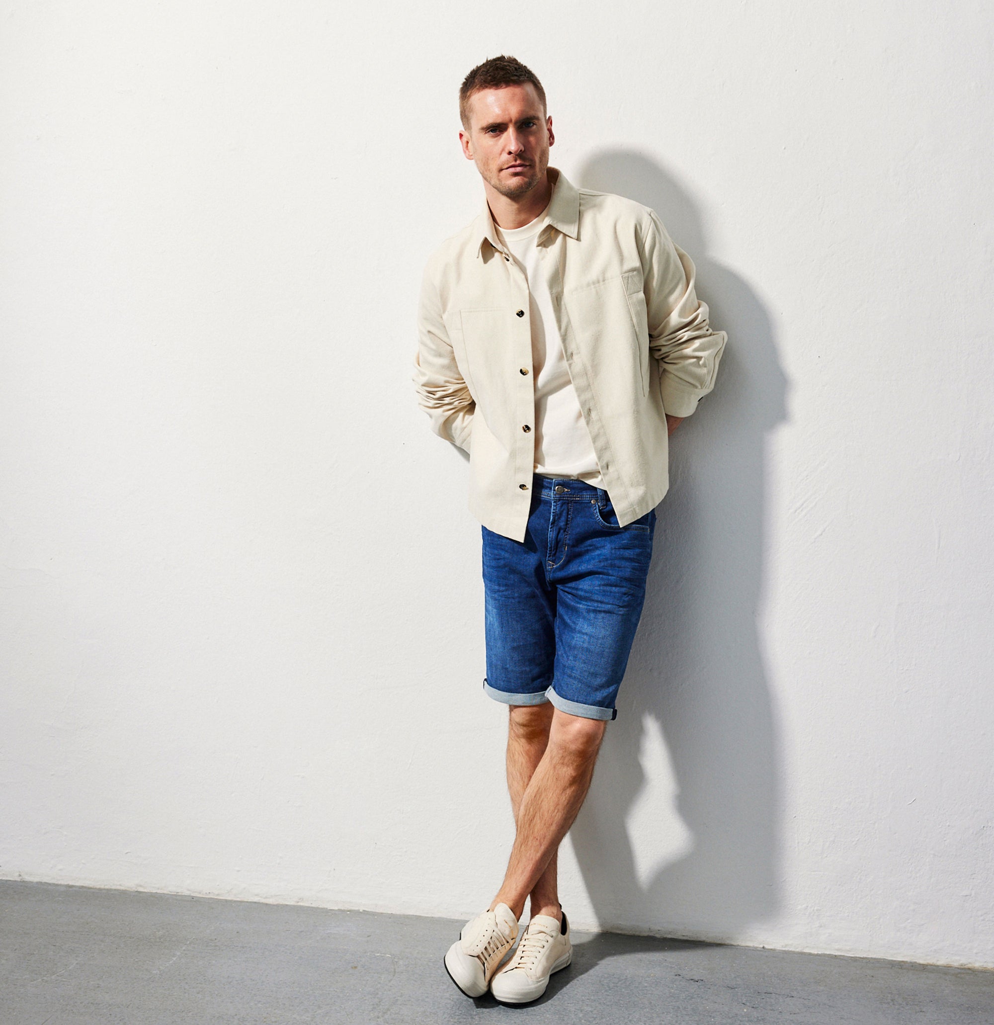 Man wearing a beige shirt, white t-shirt, blue shorts, and white sneakers against a white wall.