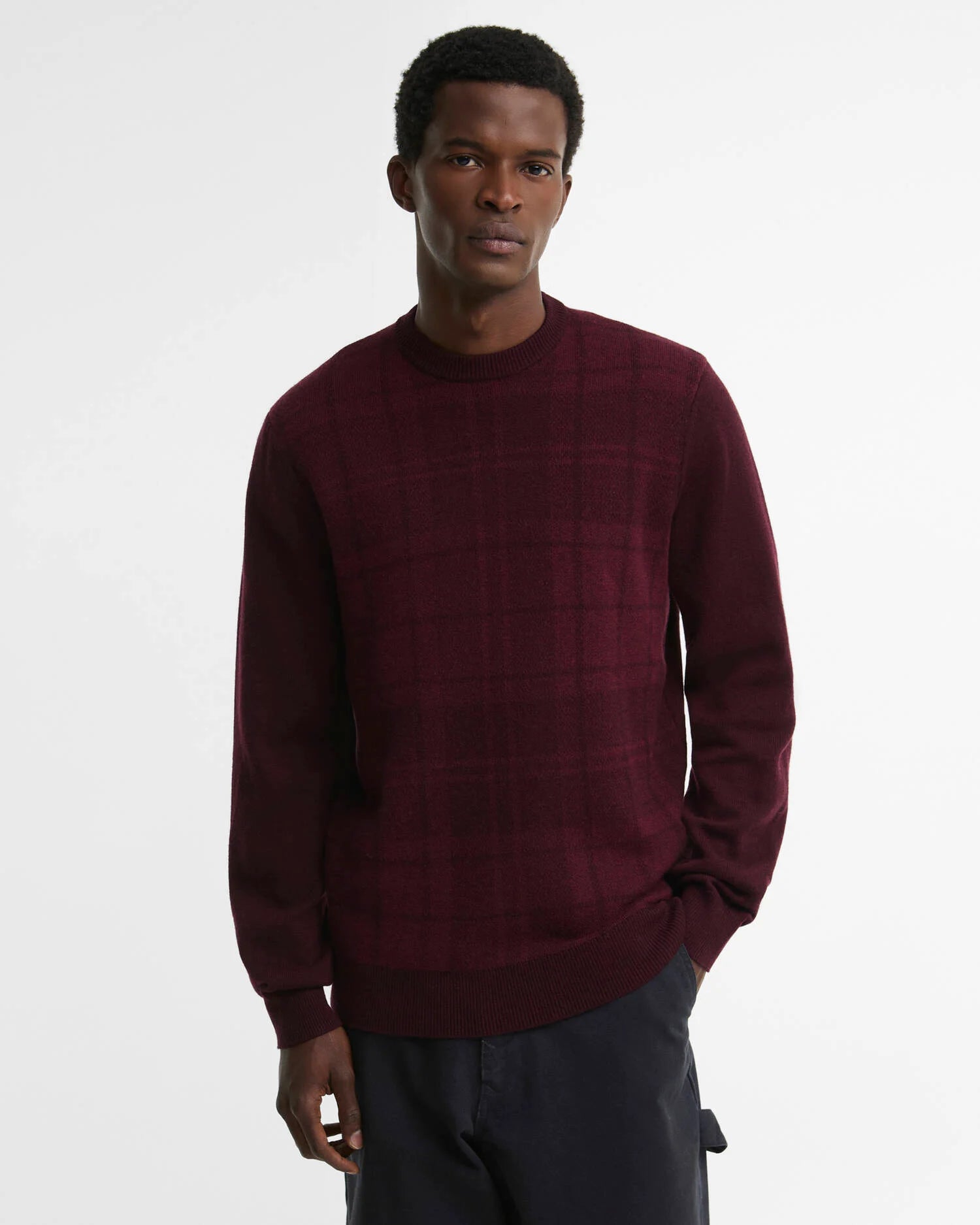 A man wearing a Barbour burgundy long-sleeve knit sweater with a subtle plaid pattern, paired with dark trousers, photographed against a clean white studio background.