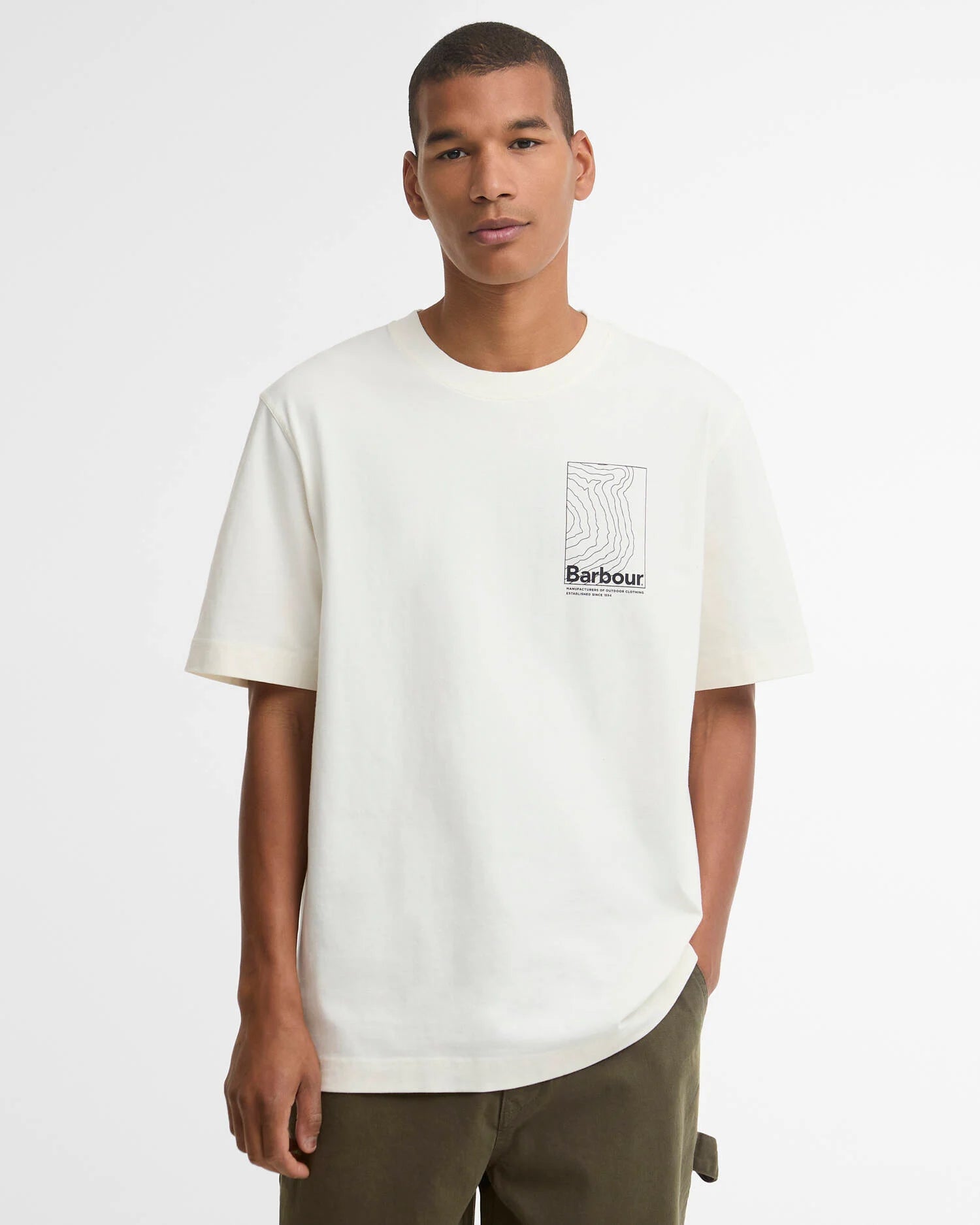 A man wearing a cream Barbour short-sleeve crewneck t-shirt with a small black Barbour logo graphic on the left chest, paired with olive green pants, against a plain white studio background.