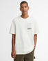 A man wearing a cream Barbour short-sleeve crewneck t-shirt with a small black Barbour logo graphic on the left chest, paired with olive green pants, against a plain white studio background.