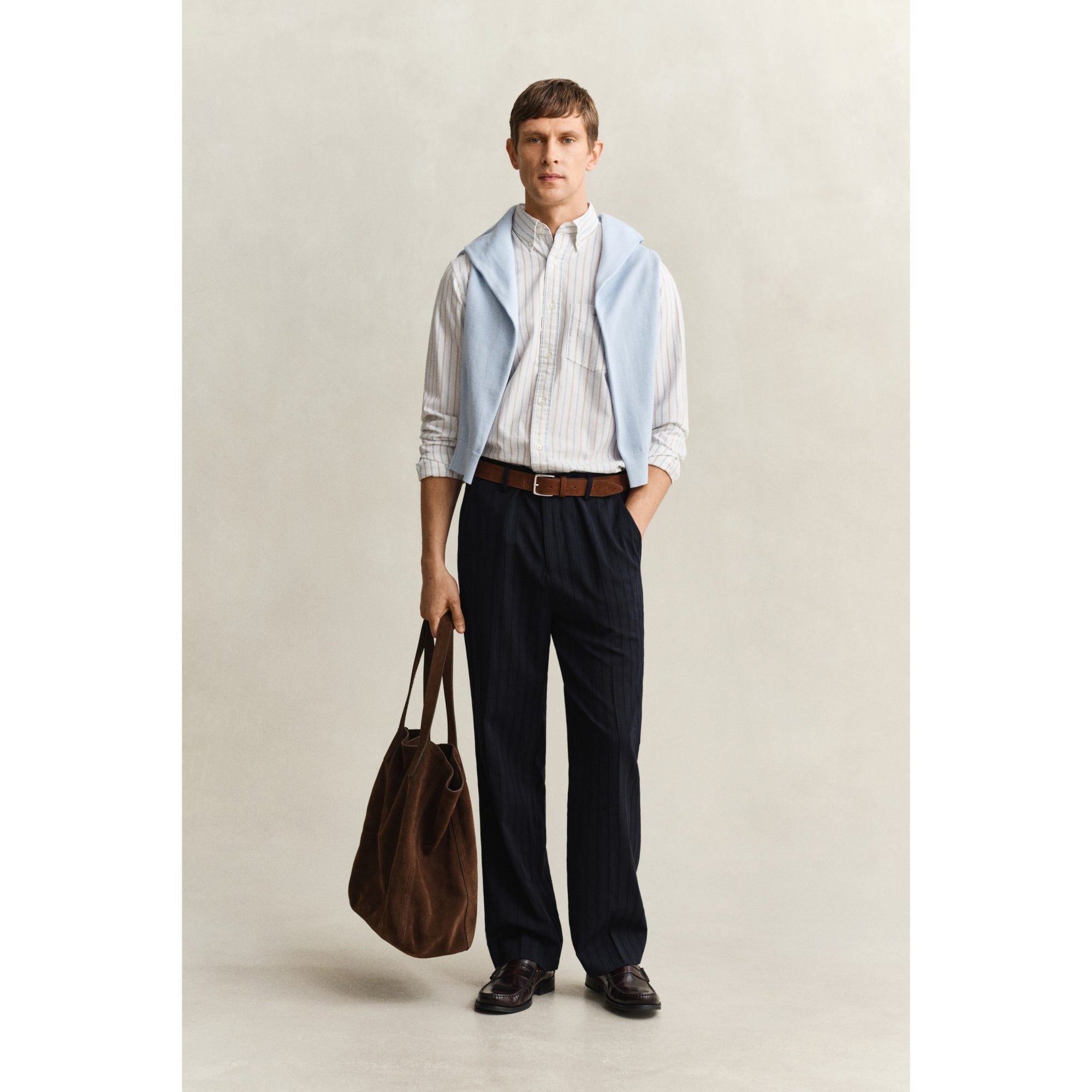 Man wearing a light blue vest over a striped shirt with dark pants, holding a brown bag on a plain background