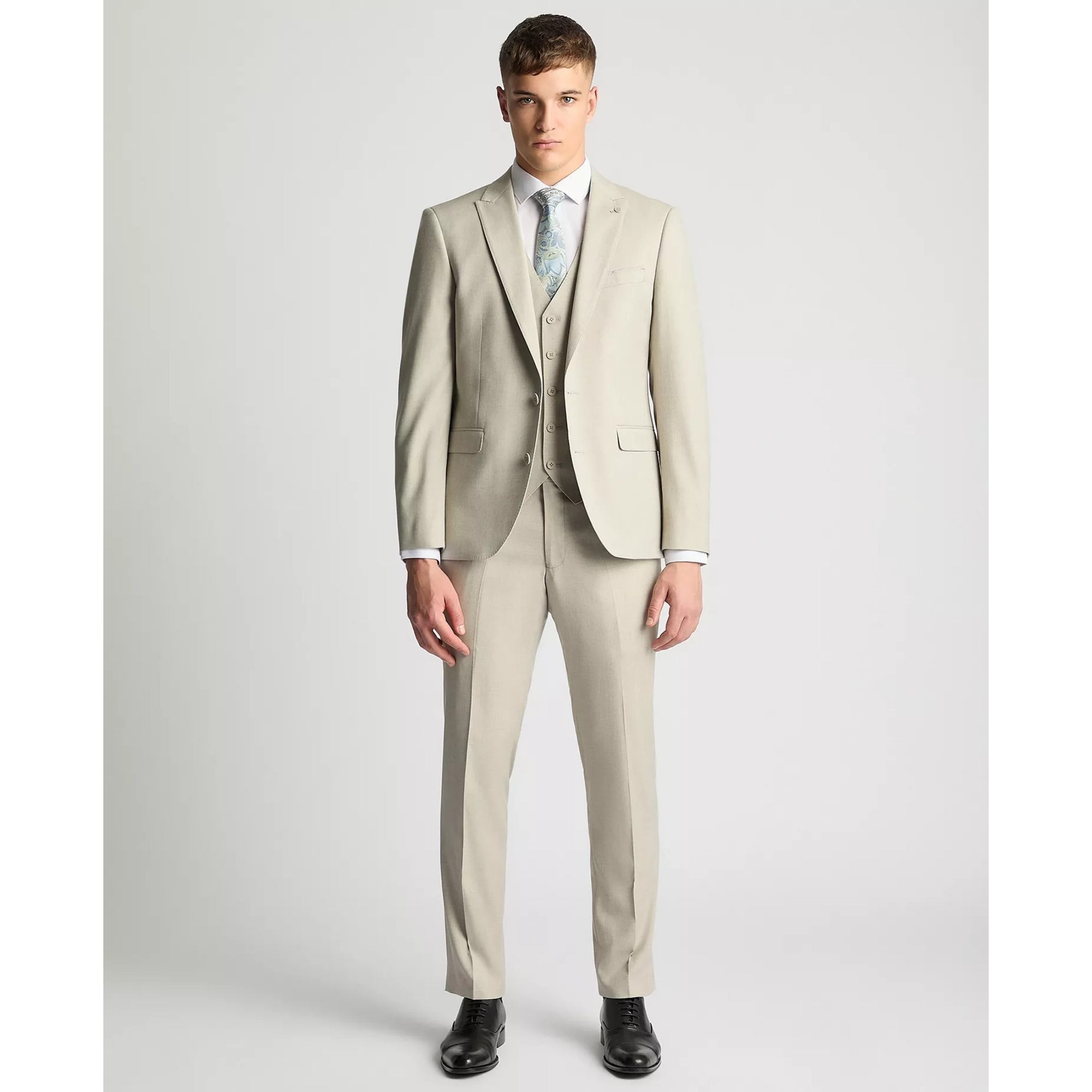 Man wearing a beige suit with a white shirt and patterned tie on a light gray background