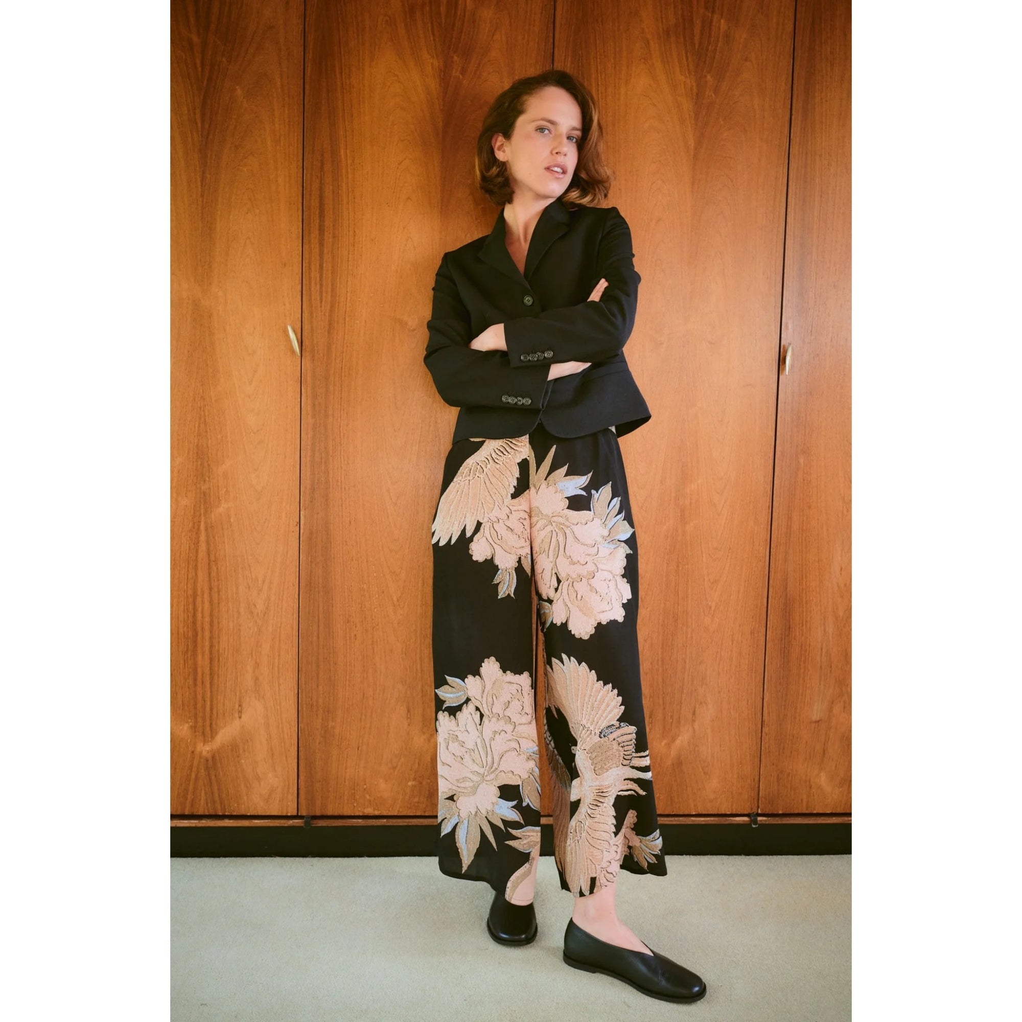 Woman wearing a black blazer and floral pants standing against a wooden wall.