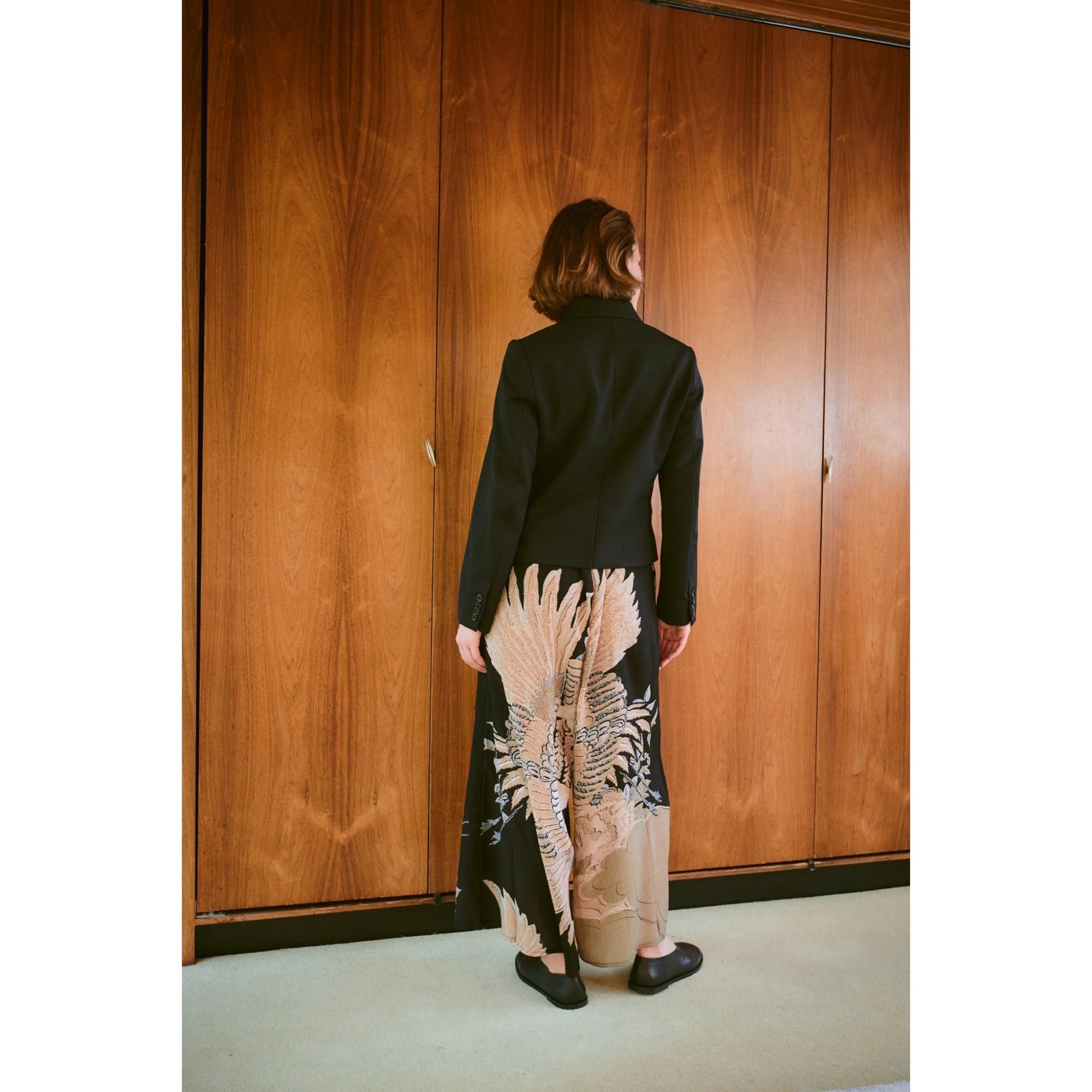 Person wearing a black jacket and patterned skirt standing in front of wooden doors.