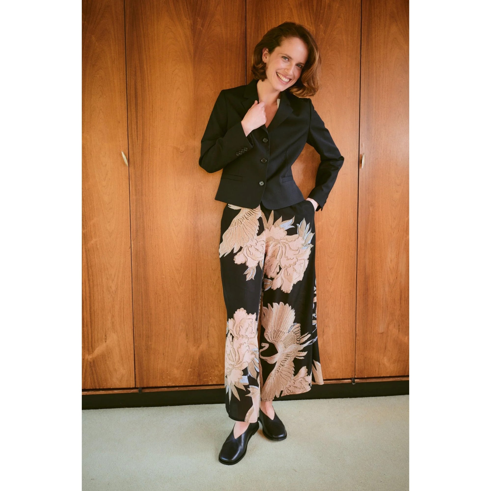 Woman wearing a black jacket and floral pants standing in front of wooden paneling.