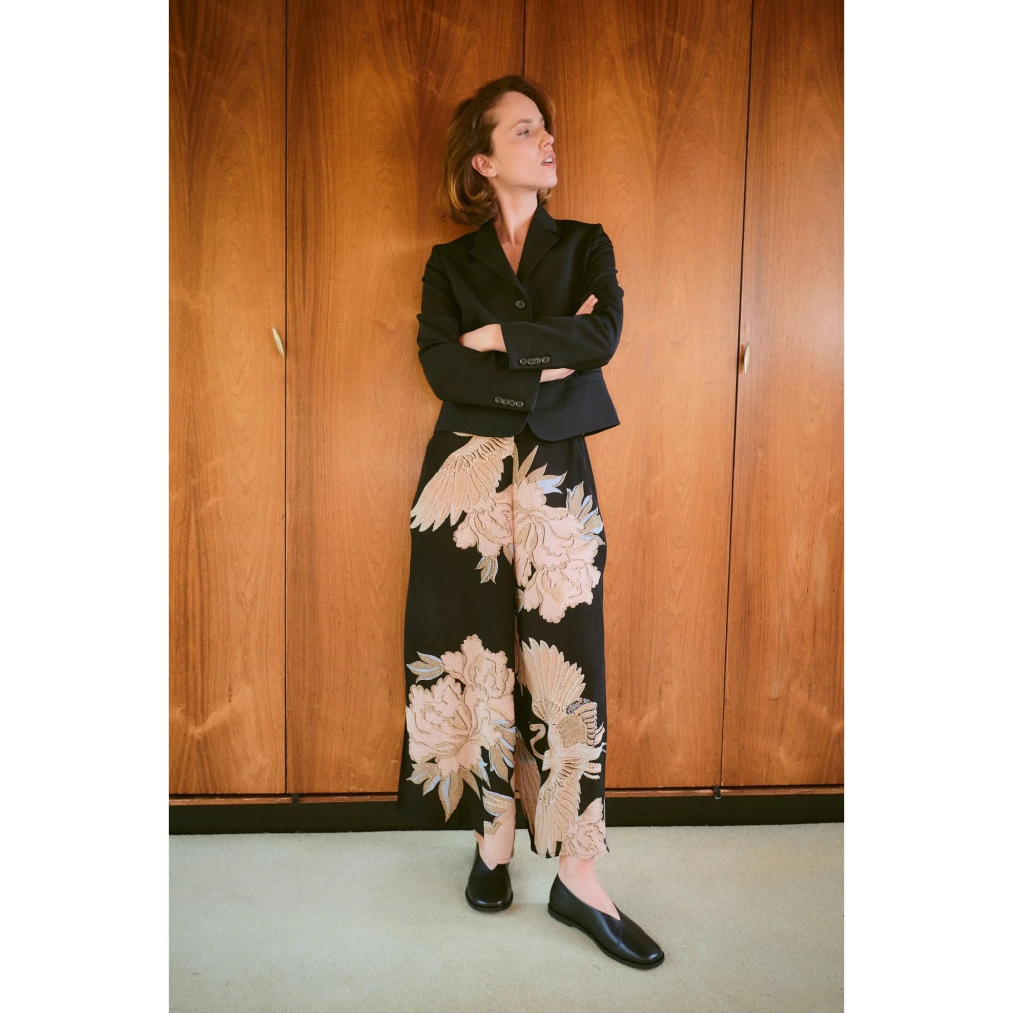 Woman wearing a black blazer and floral pants standing against a wooden wall.
