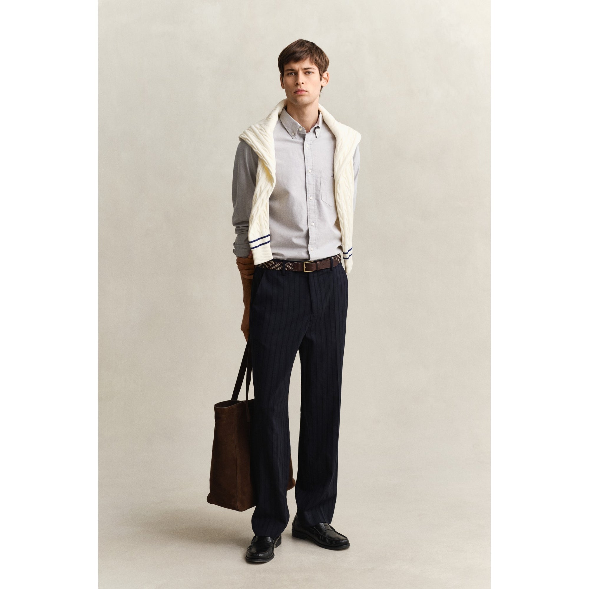 Man wearing a light gray sweater, dark pants, and holding a brown bag against a plain background