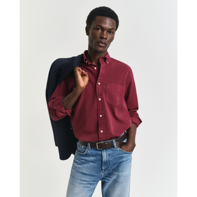 Man wearing a GANT red shirt and blue jeans with a dark jacket over his shoulder on a light gray background