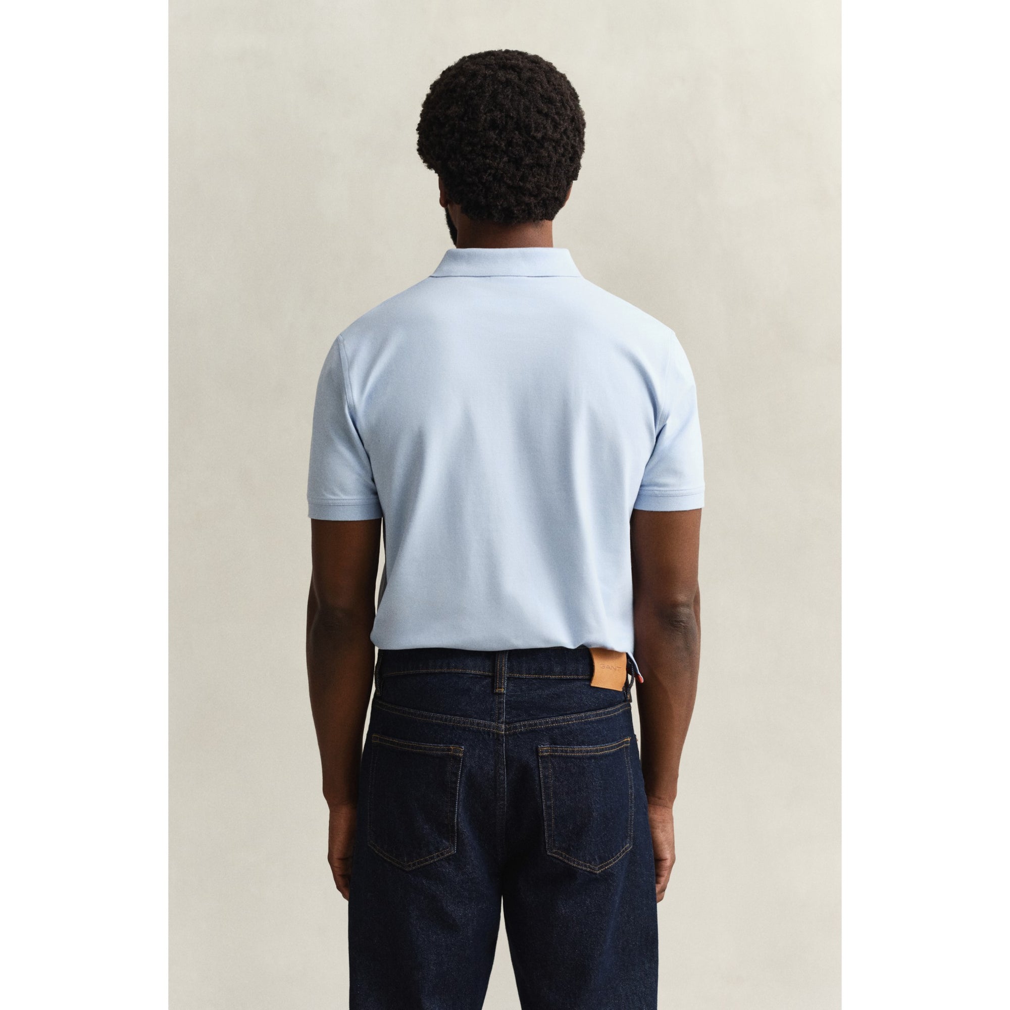 Person wearing a light blue polo shirt and dark jeans against a beige background