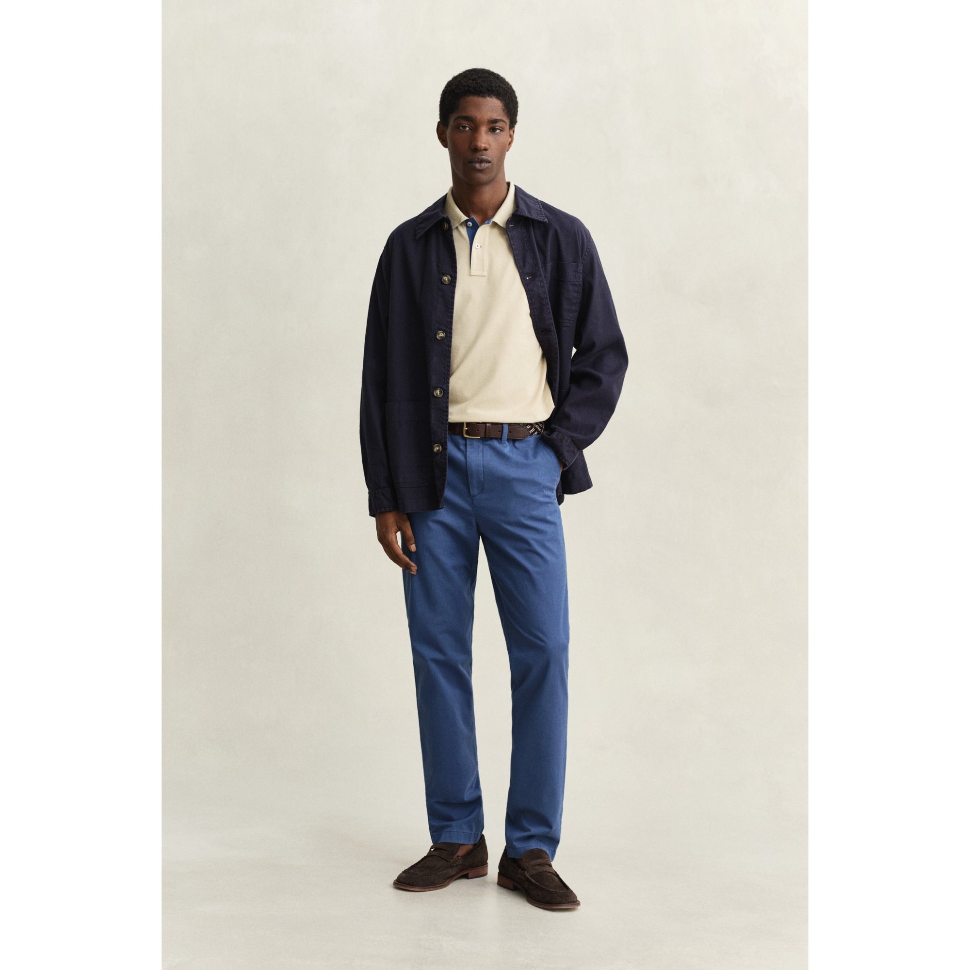 Man wearing a navy jacket, beige shirt, blue pants, and brown shoes on a plain background