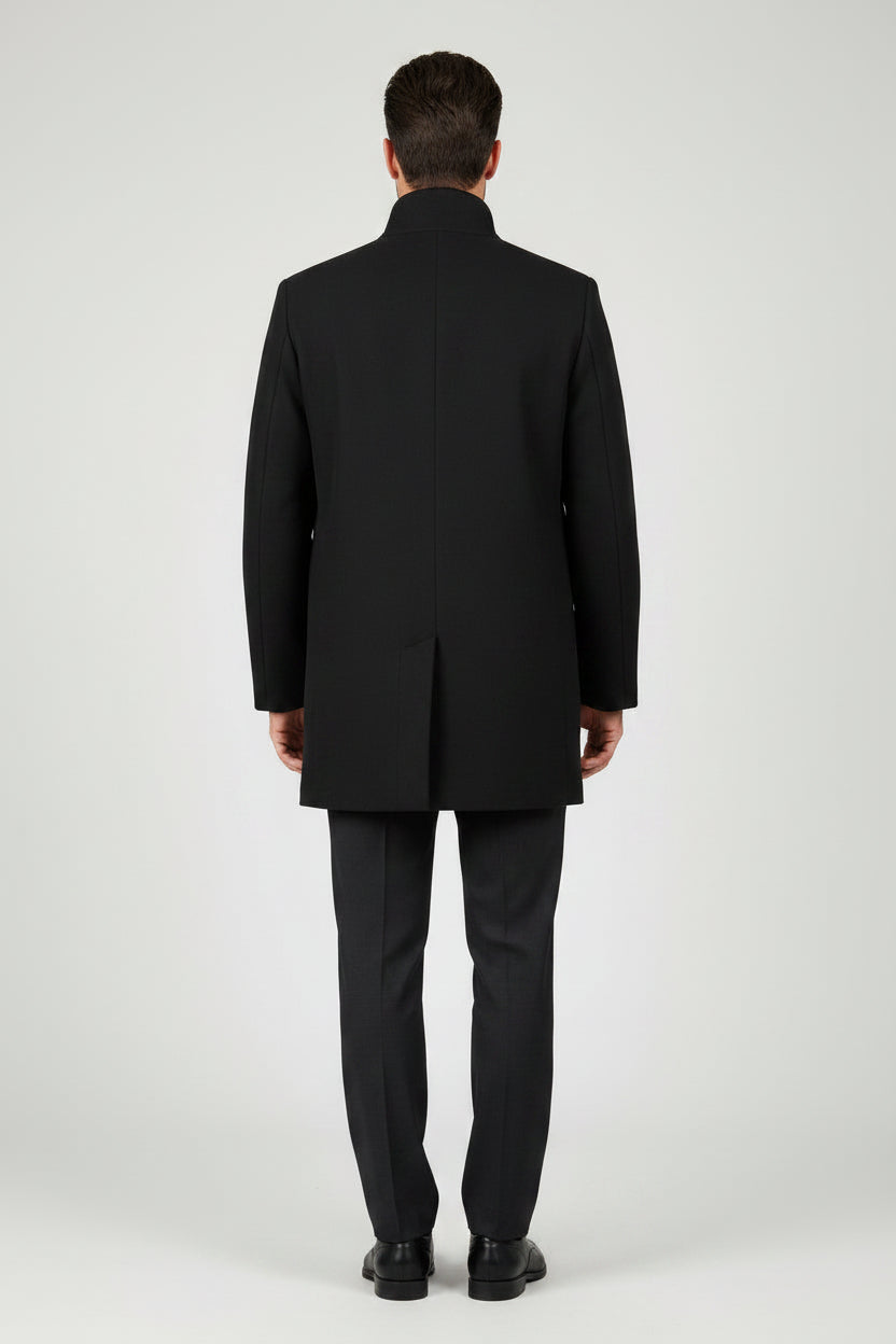 A man modeling a black mid-length coat with a stand collar, three front buttons, and front pockets, paired with black trousers and black dress shoes, against a light gray studio background.