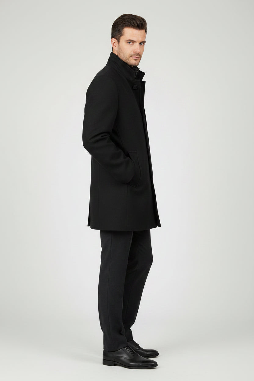 A man modeling a black mid-length coat over a black turtleneck sweater, black trousers, and black dress shoes, standing against a light gray studio background.