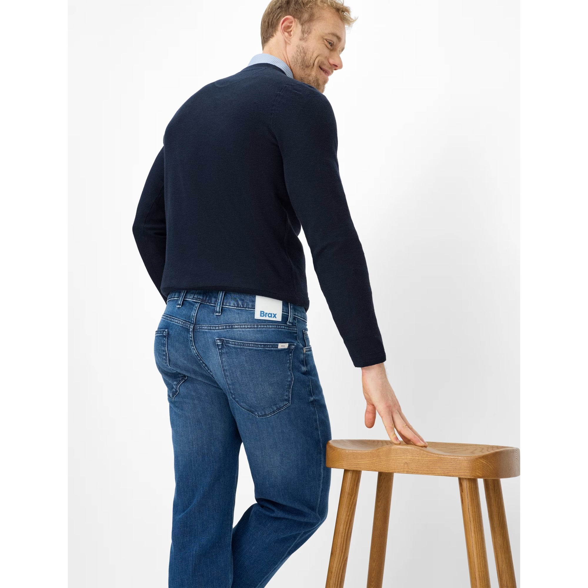 Man wearing a navy sweater and blue jeans standing next to a wooden stool on a white background