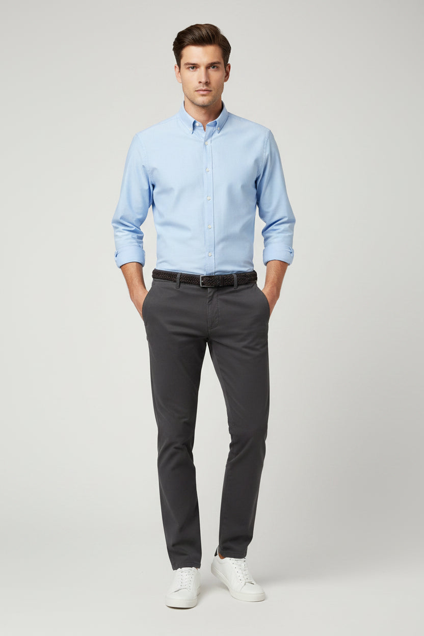 A man modeling a light blue long-sleeve button-down shirt, dark gray trousers, a black belt, and white sneakers against a light gray studio background.
