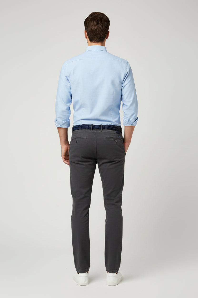 A man modeling a light blue long-sleeve button-down shirt, dark gray trousers, a black belt, and white sneakers against a light gray studio background.