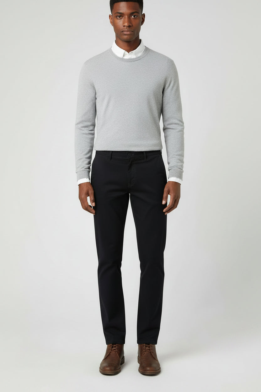 A man wearing a light gray long-sleeve knit sweater over a white collared shirt, paired with dark trousers and brown leather shoes, standing against a light gray studio background.
