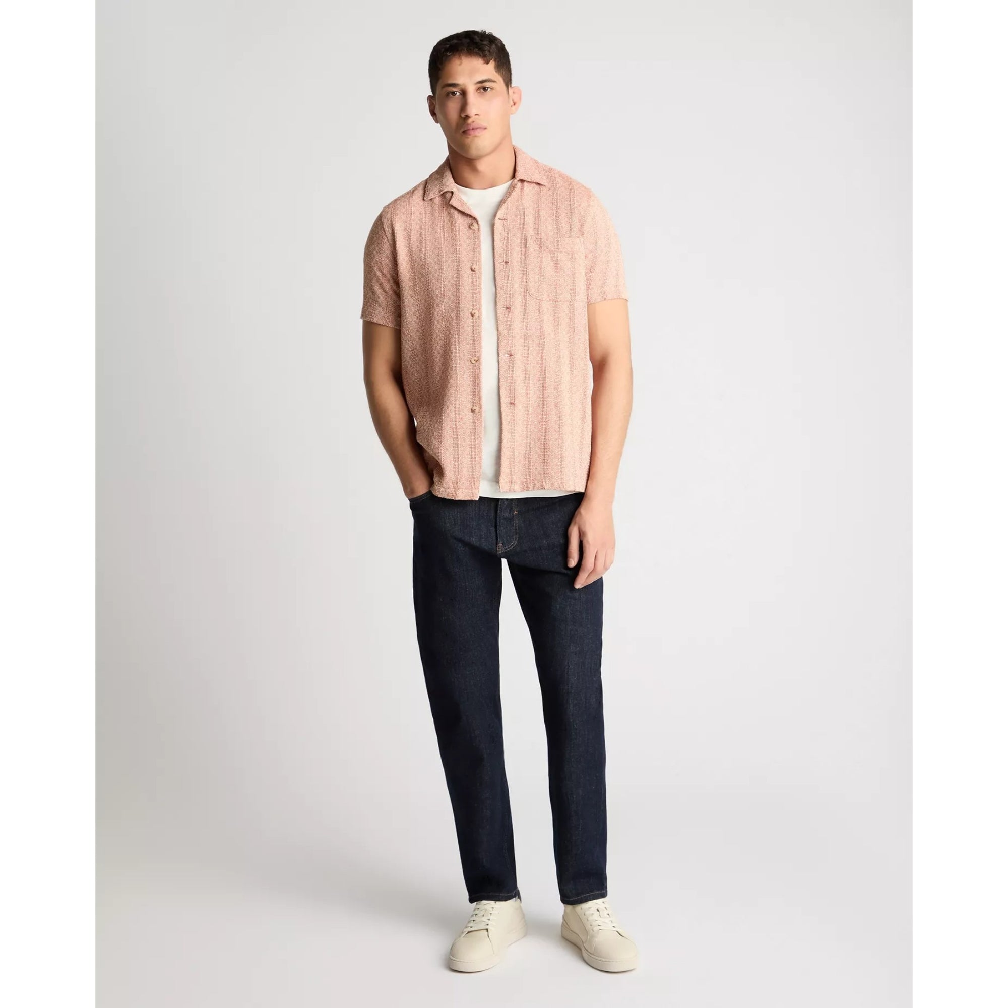 Man wearing a light pink button-up shirt over a white t-shirt, dark jeans, and white sneakers on a plain background