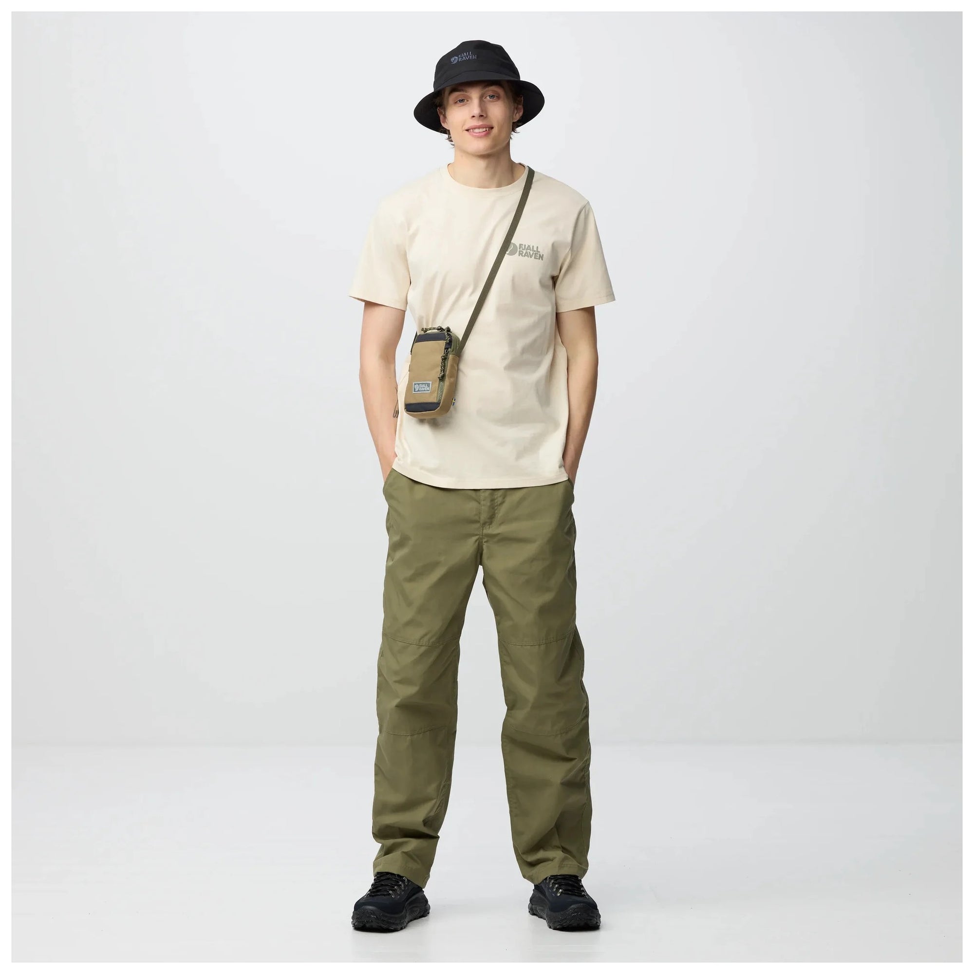 Person wearing a beige t-shirt, green pants, black shoes, and a black hat with a crossbody bag on a white background