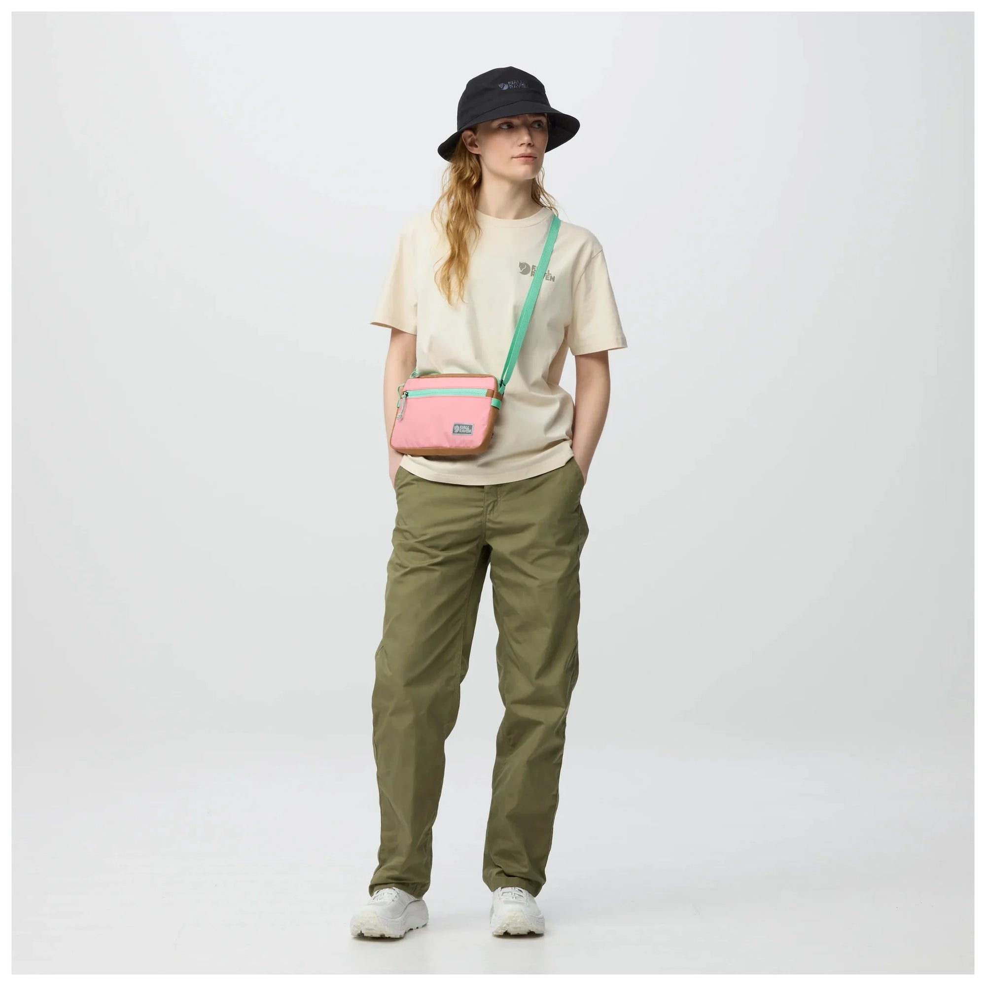 Person wearing a beige shirt, green pants, and a black hat, carrying a pink crossbody bag on a white background.