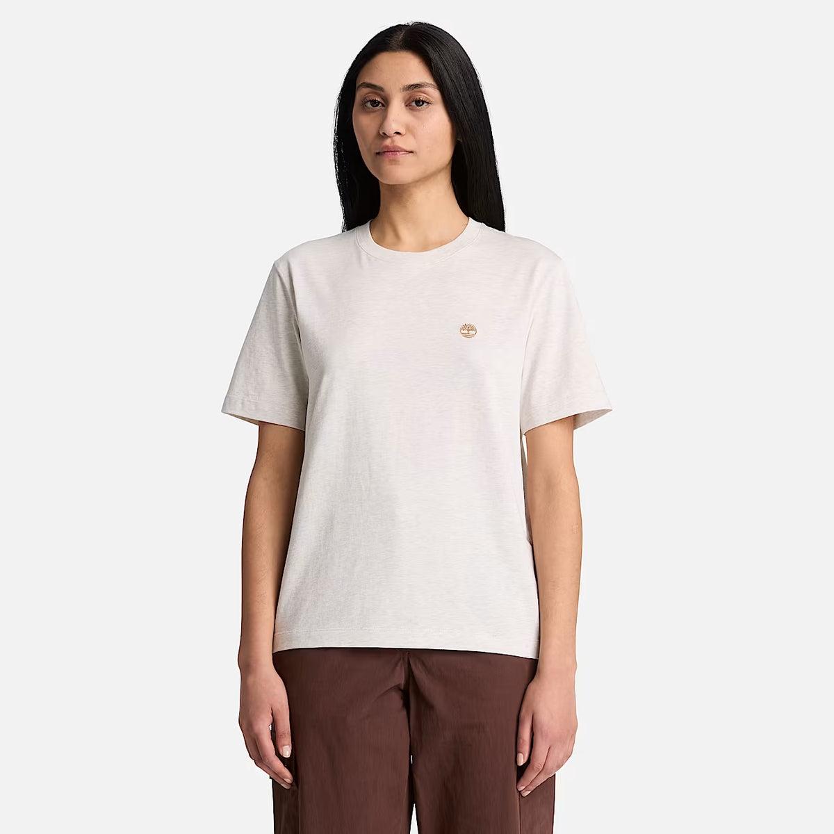 A woman modeling a short-sleeve crewneck t-shirt in a light oatmeal beige color with a small gold embroidered logo on the left chest, paired with brown pants, against a solid light gray background.