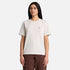 A woman modeling a short-sleeve crewneck t-shirt in a light oatmeal beige color with a small gold embroidered logo on the left chest, paired with brown pants, against a solid light gray background.